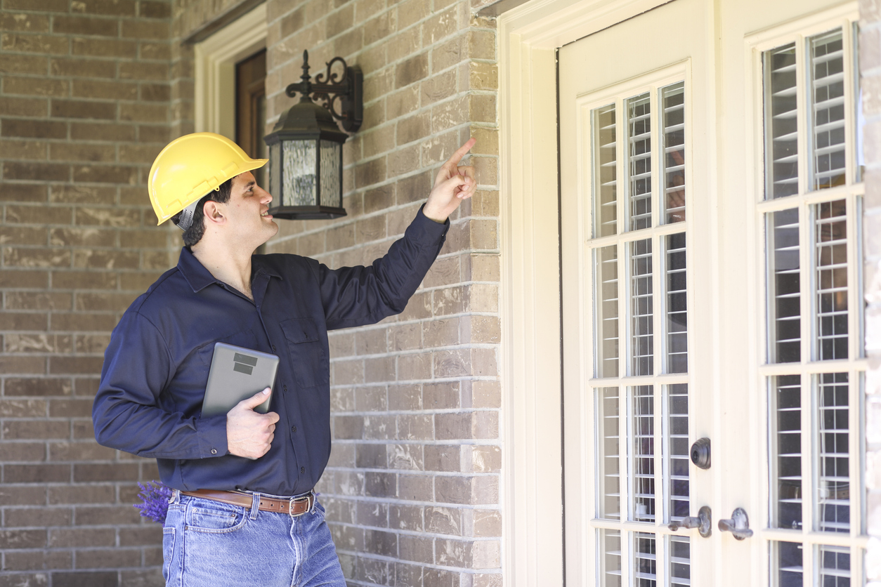 Latin descent man inspects home exterior. He could be a home inspector, insurance adjuster, exterminator or a variety of blue collar occupations. He holds a digital tablet, wears hard hat and blue work shirt.