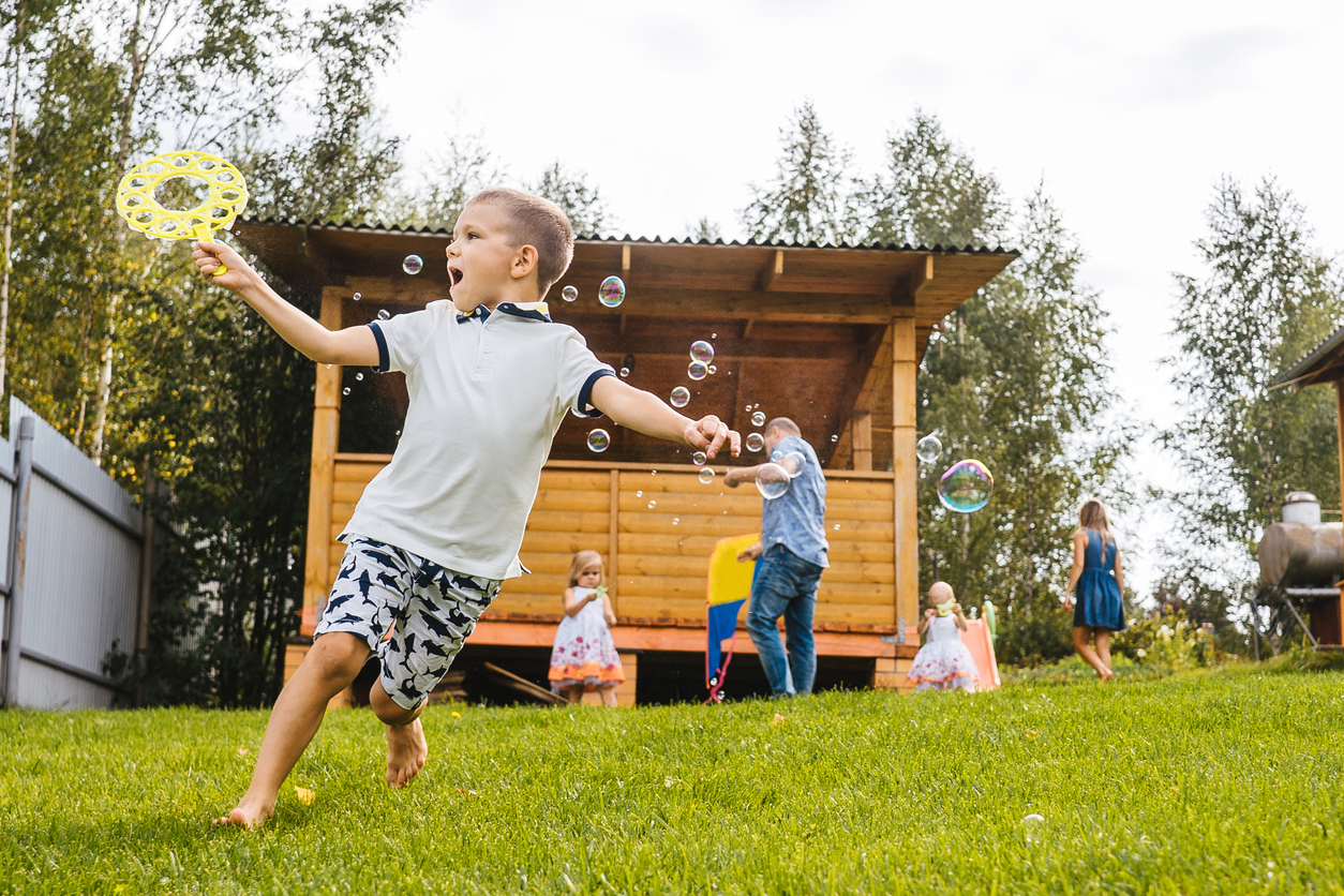 Little boy run with blowing soap bubbles. Backyard, family on background.