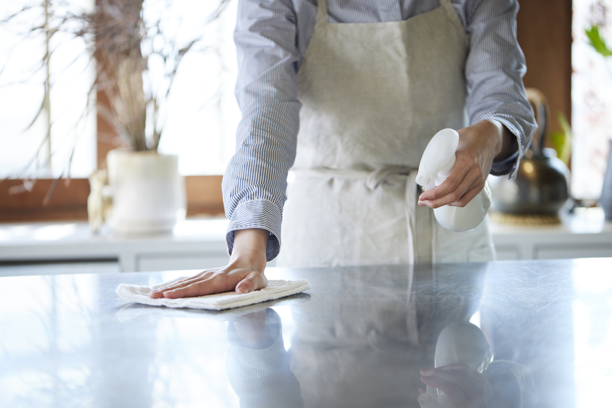 a woman wearing an apron uses a spray bottle and rag to wash a stainless steel counter top.