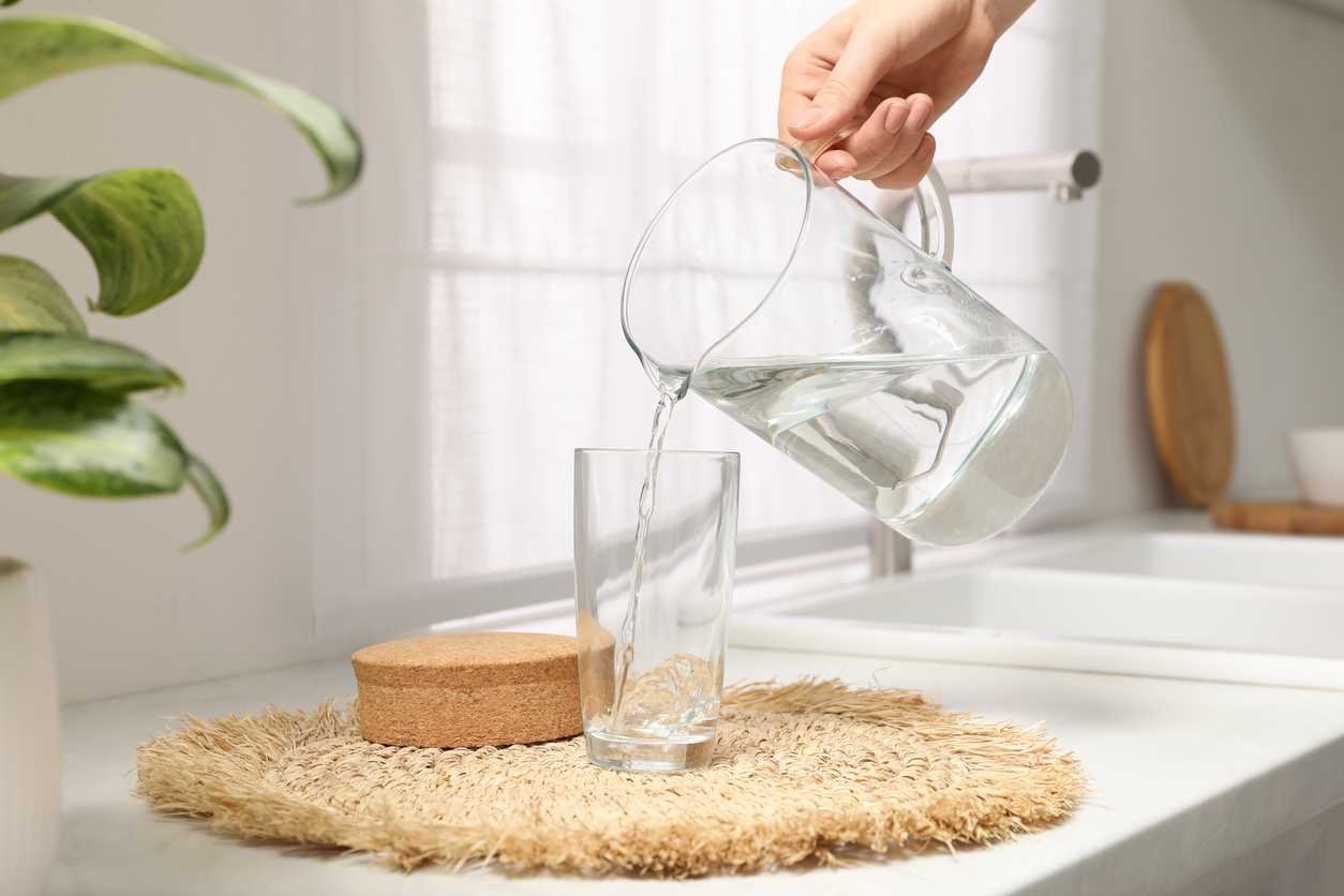 Woman pouring water into glass from glass jug on countertop in kitchen.