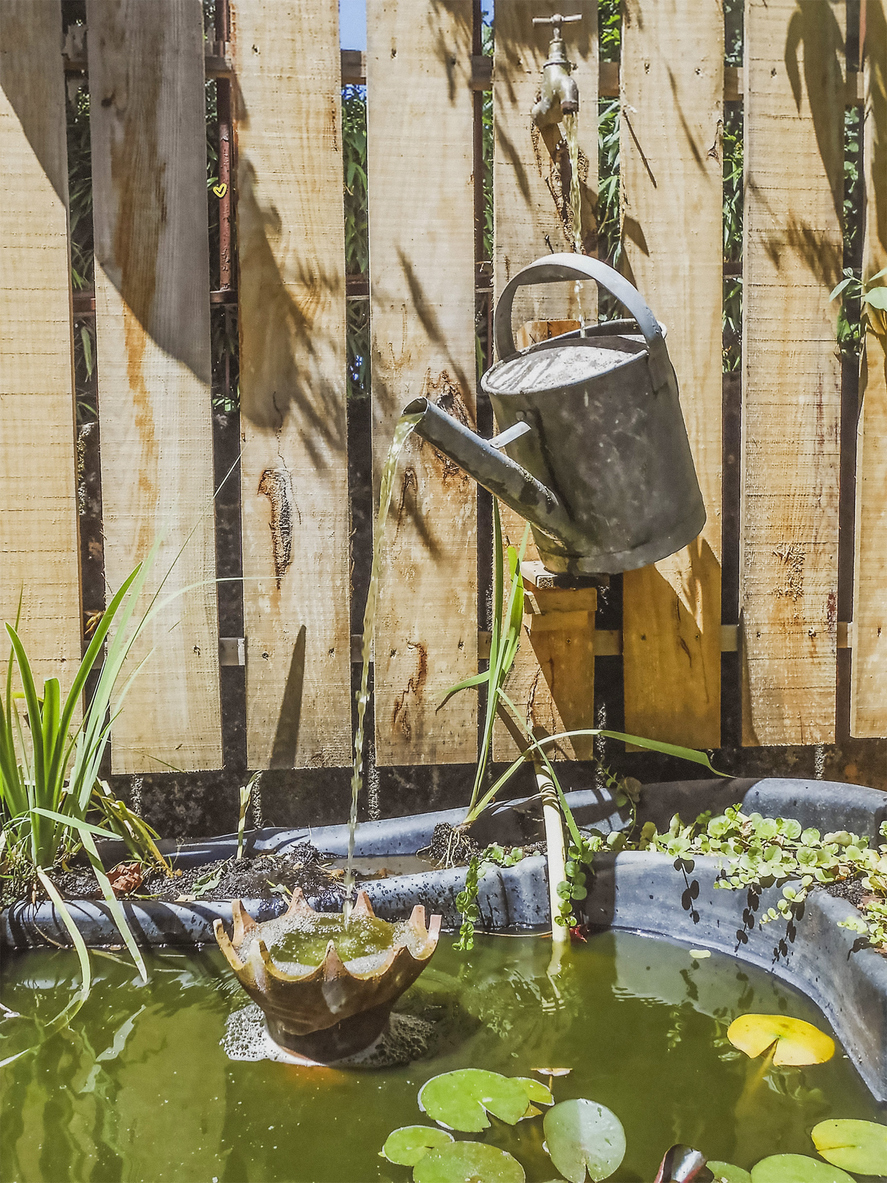 Water pours from a mounted watering can into a fountain in a backyard.