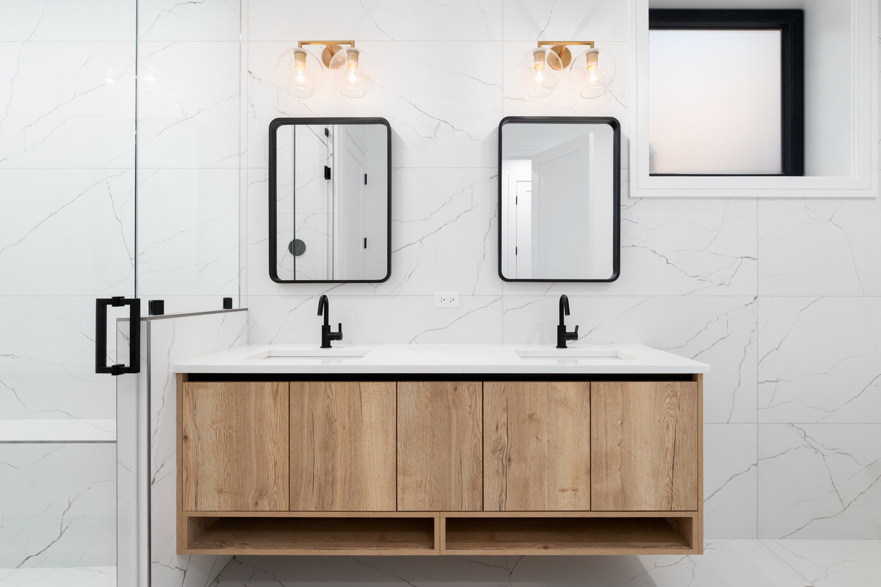 A beautiful bathroom with marble walls, a floating natural wood cabinet, marble countertop, and gold lights mounted above the black mirrors.