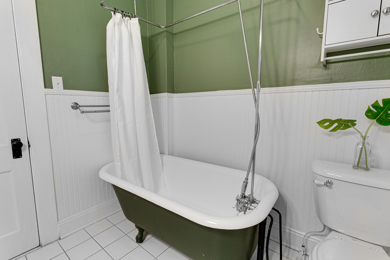 Simple and elegant bathroom with green walls, white bathtub, shower curtain, wall-mounted cabinet, glass vase with monstera leaves, toilet with flower, and modern design.