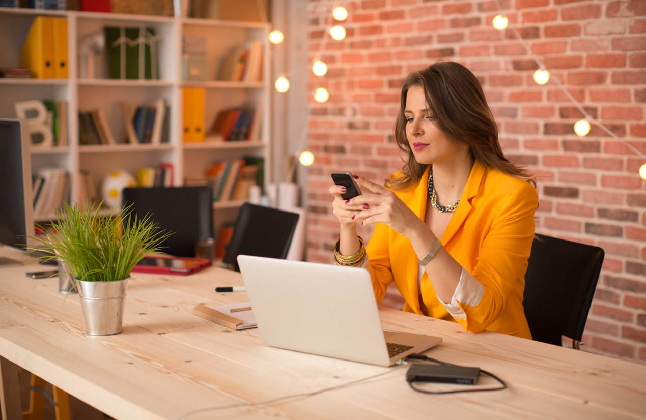 A-string-of-lights-hangs-on-a-brick-wall-behind-a-woman-sitting-at-a-desk-and-looking-at-her-phone.