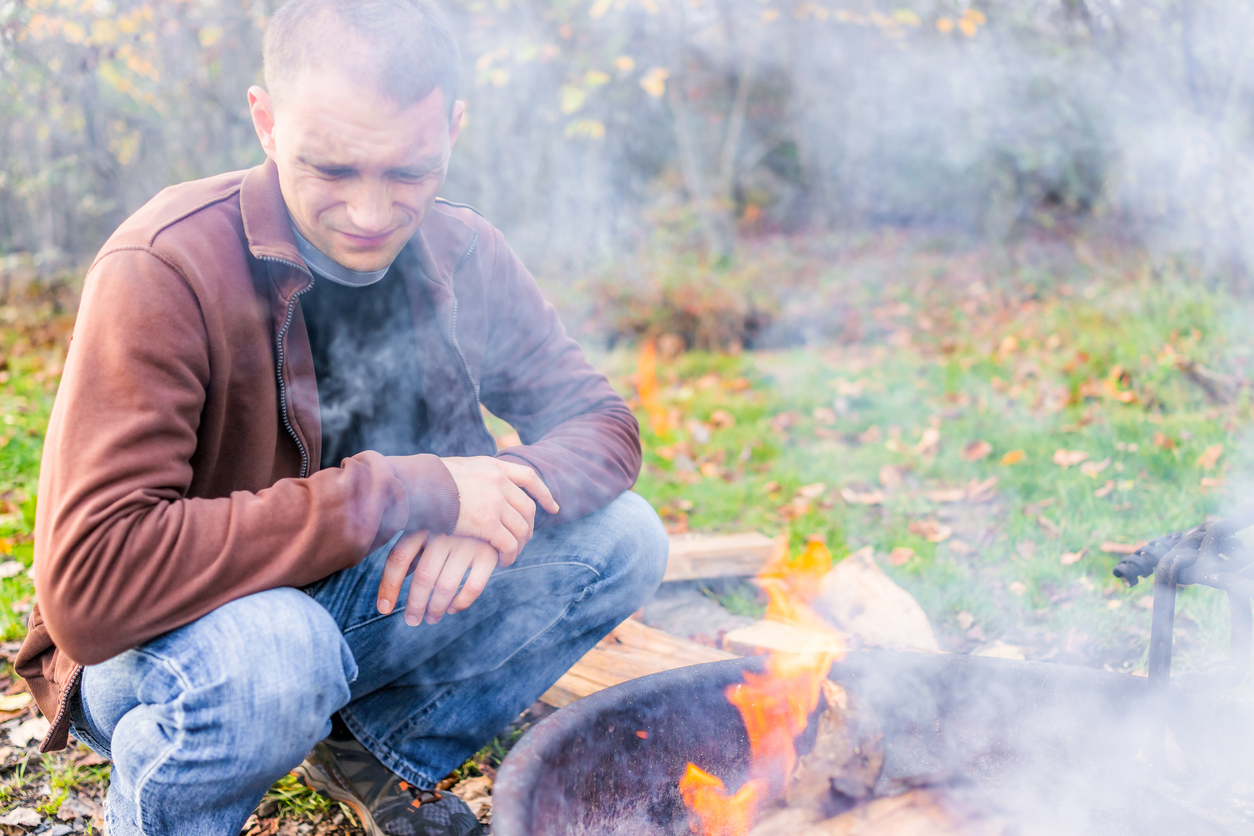 man-sits-next-to-a-fier-pit-and-grimaces-at-the-smoke