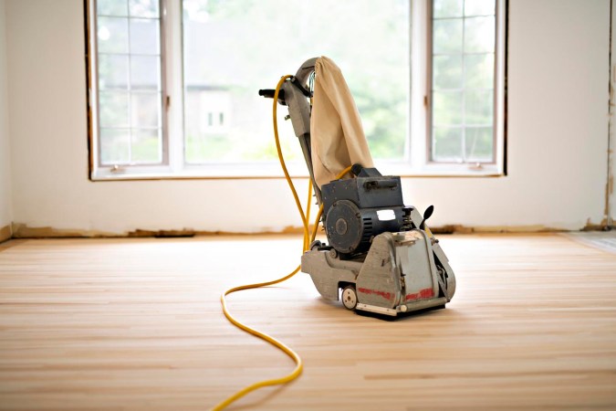 A floor sander sits on an unfinished wood floor in an empty room.