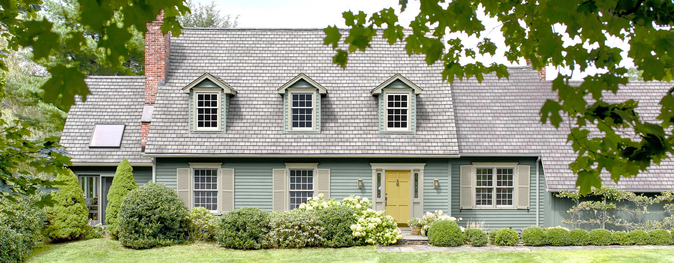 A two story home with dormer windows is painted a pale blue with a yellow door and greenery surrounding.