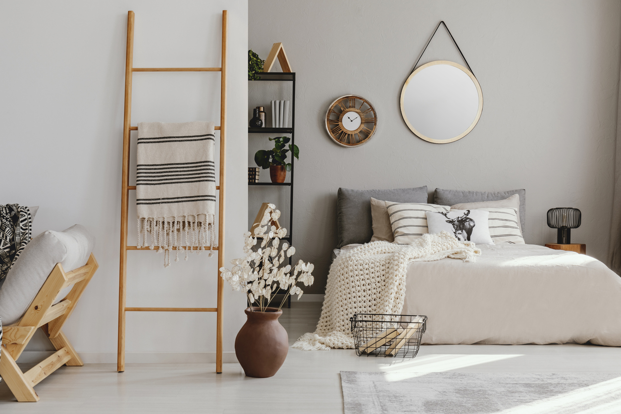 An open space bedroom interior with double bed with knit blanket and many pillows, a rack with books and decor, a carpet on the floor and a blanket ladder leaning on the wall outside