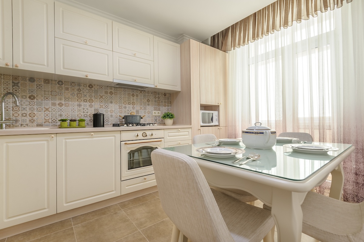 A large white kitchen with a large window covered by sheer cutrains.