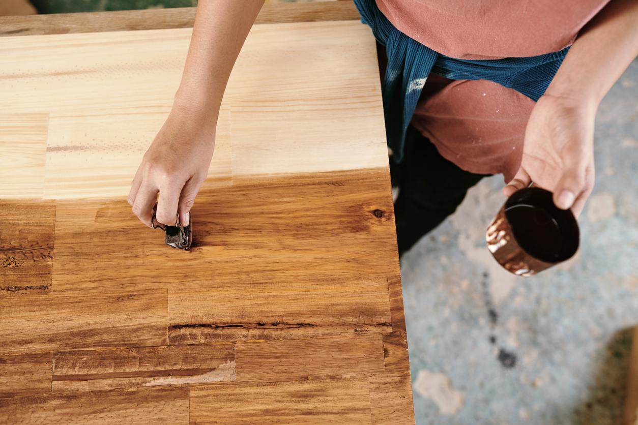 A carpenter's hands apply a wood varnish onto a wooden butcher block using a sponge. 