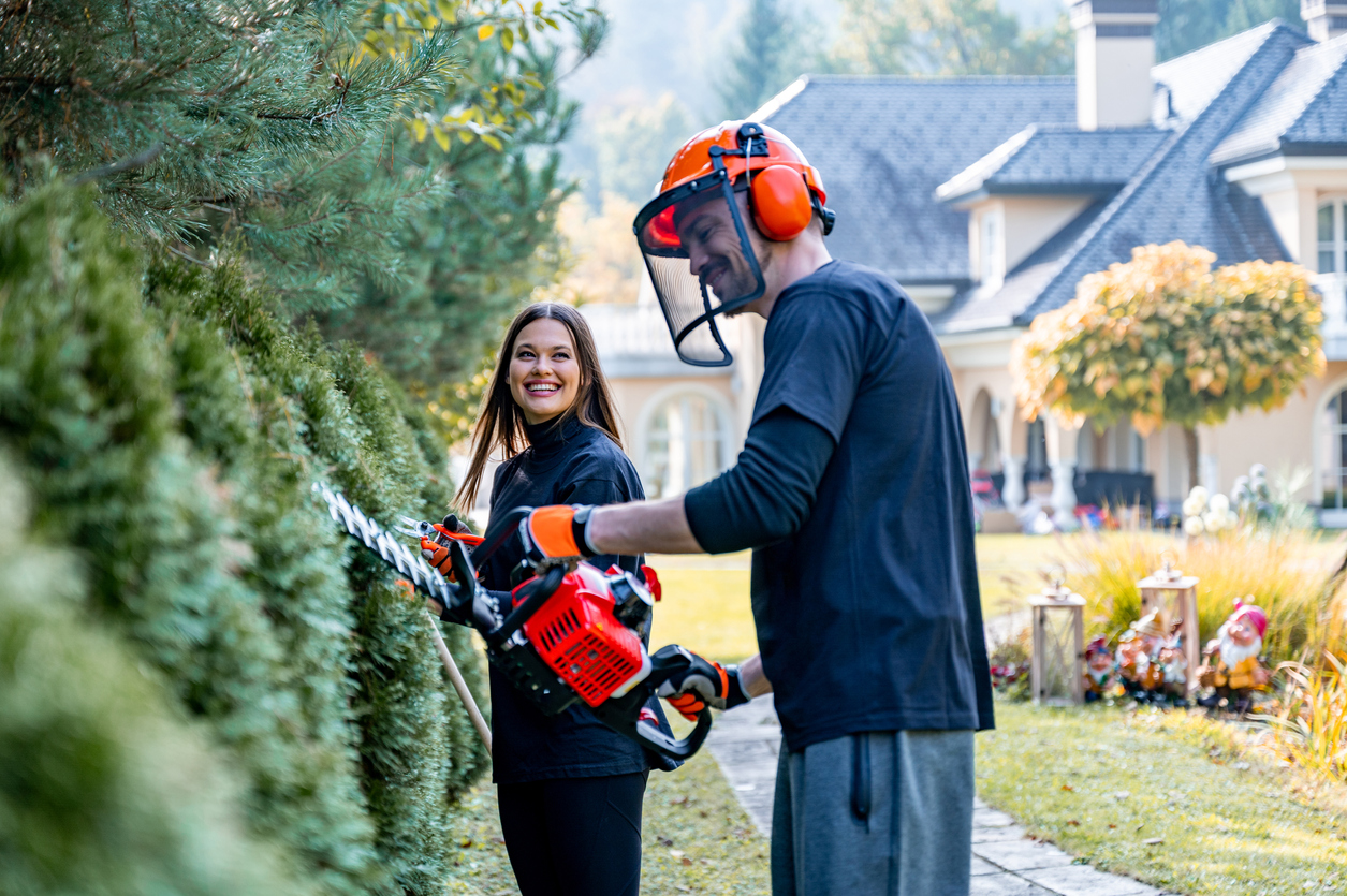 Young adult couple trimming a hedge with power saw and hedge clippers.