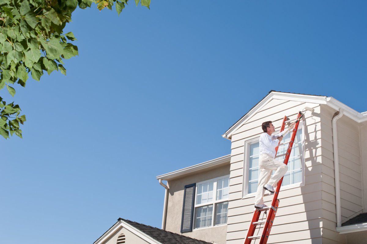 A-person-stands-on-an-extendable-ladder-and-paints-the-second-story-of-a-house-in-off-white.