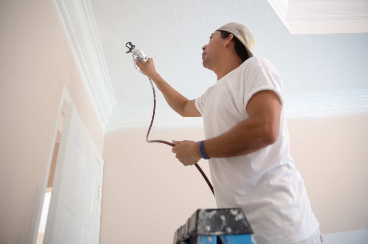 House painter using a spray paint gun to paint the crown molding on the ceiling in home, standing on a ladder. He's wearing a white t shirt and painters pants. He is painting over the door to the room.