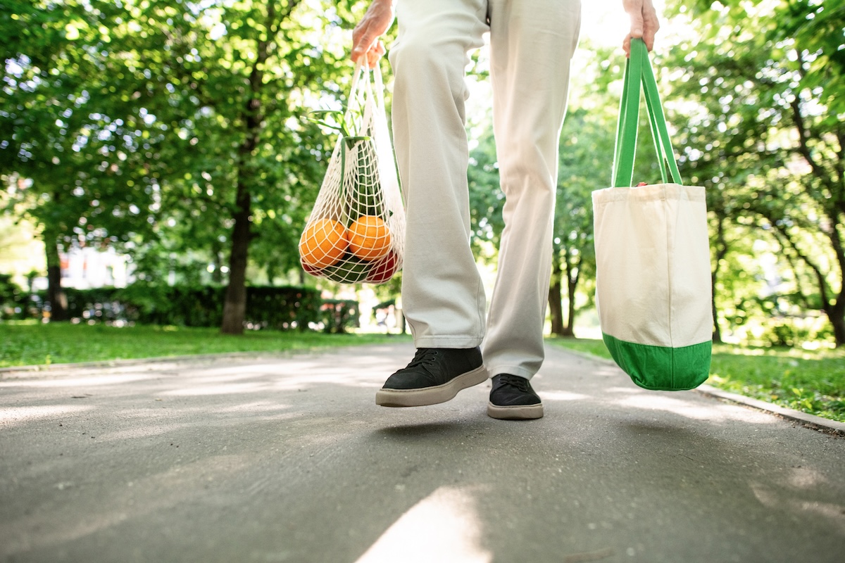 A man in white pants carries tote bags with groceries.