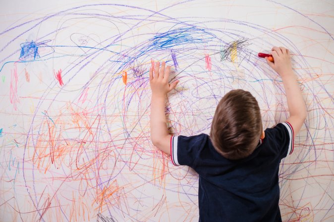 Little boy with brown hair and navy blue shirt colors white wall with crayon.