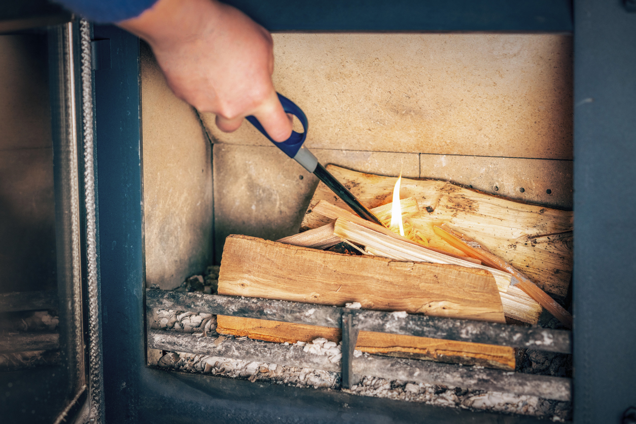 A-hand-uses-a-long-handle-lighter-to-ignite-wood-in-a-wood-burning-stove-on-top-of-existing-ashes.