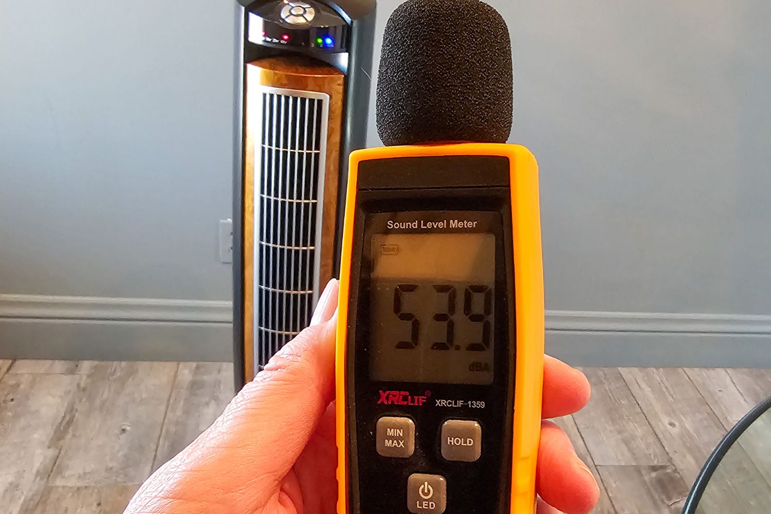 A person standing near the Lasko Wind Curve tower fan while holding a decibel meter that reads 53.9 decibels.