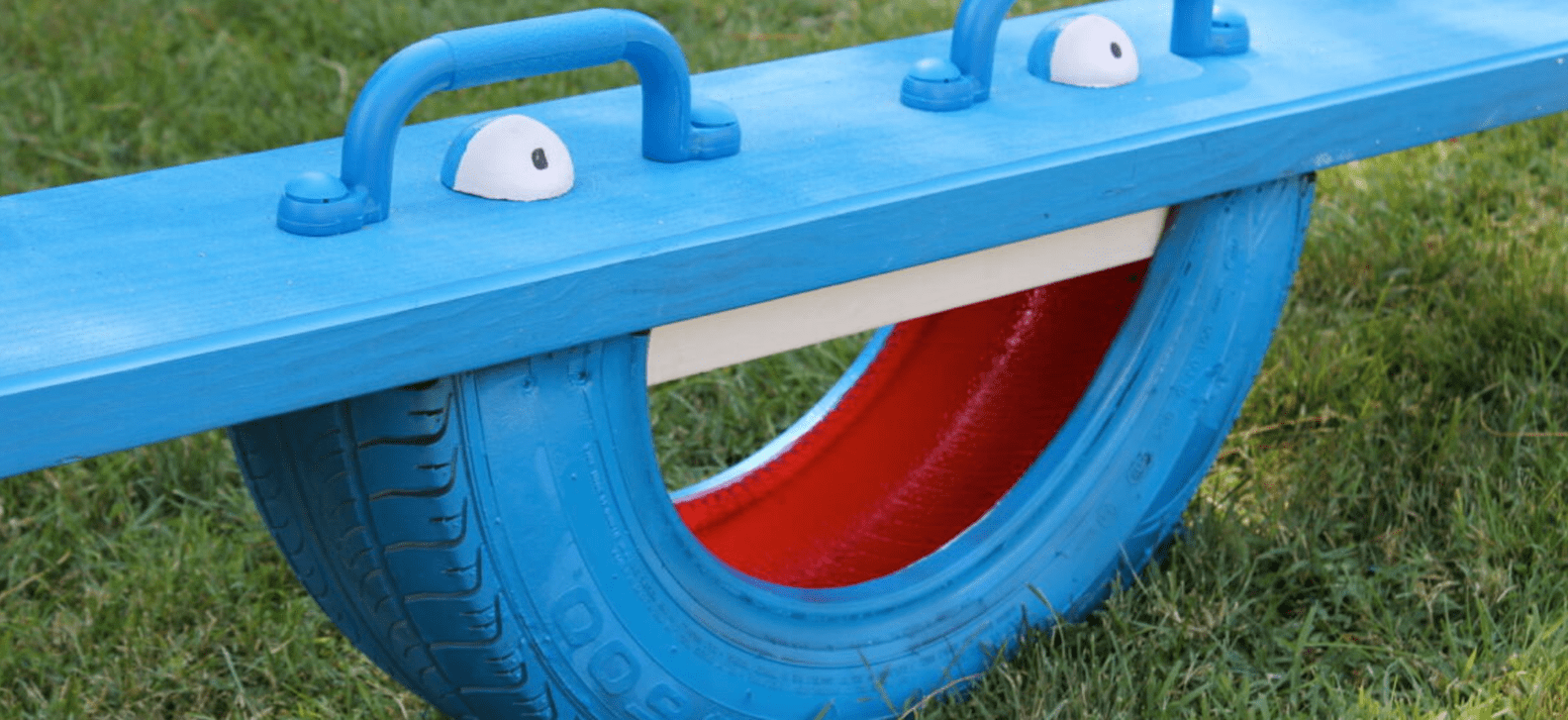 Close view of a seesaw made from an old tire painted blue with a cartoon face and blue plank on top