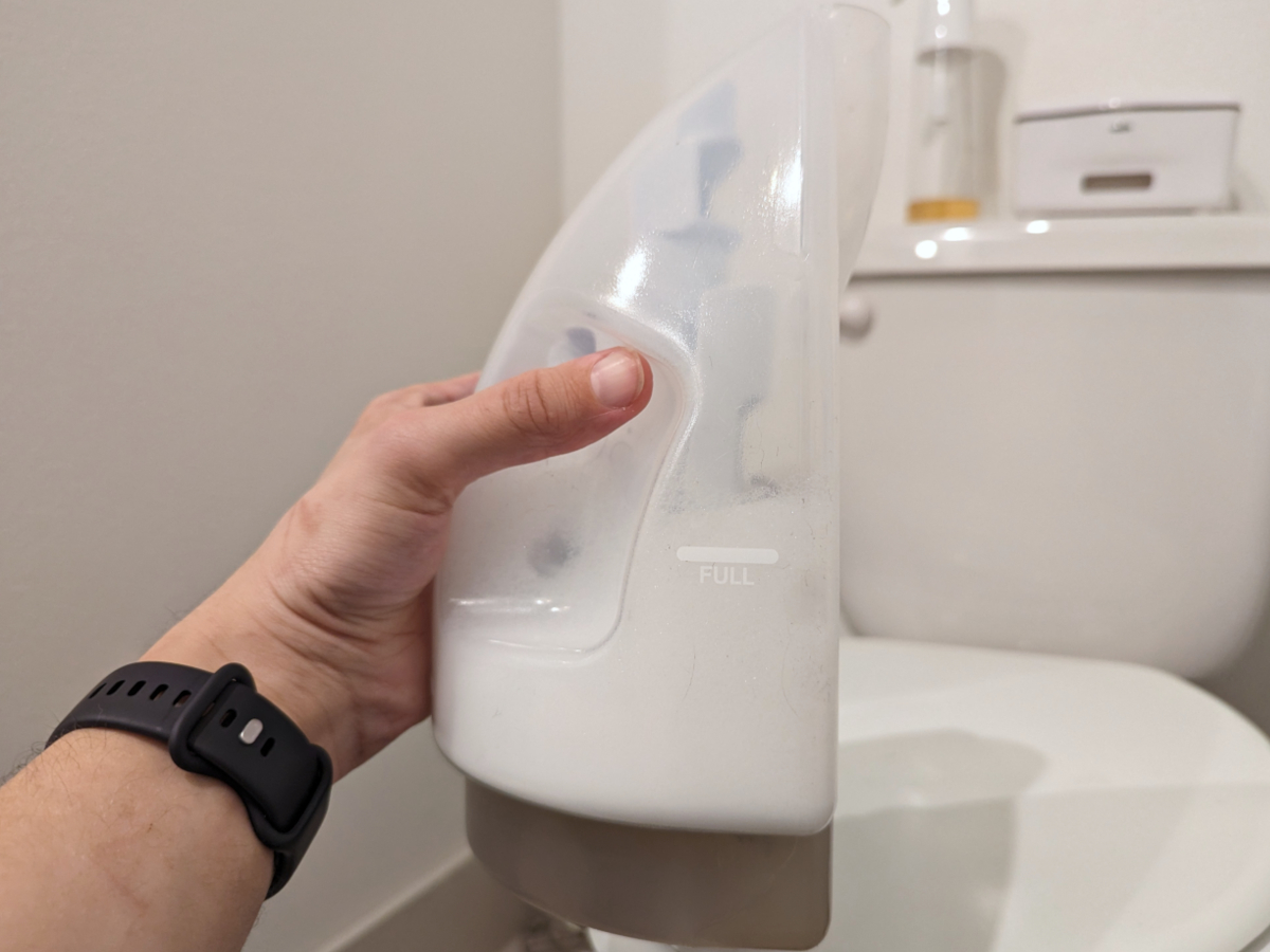 A man holding the semi-full dirty water tank of a Bissell Little Green carpet cleaner in front of a closed toilet.