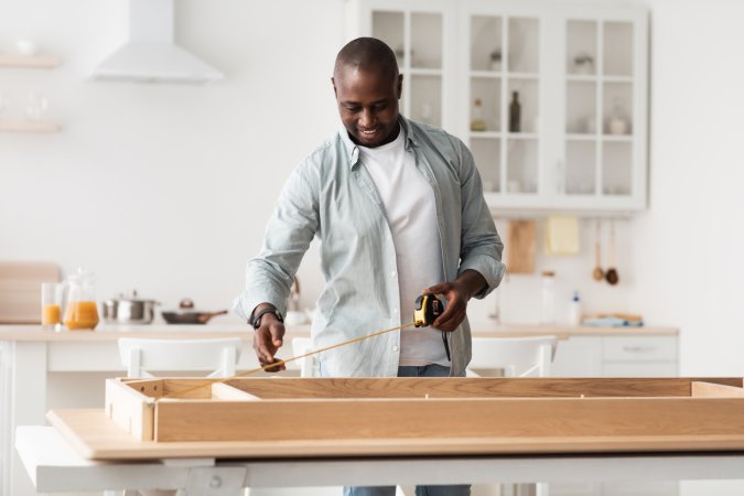 man measuring table with roulette, assembling desk in kitchen interior with various tools.