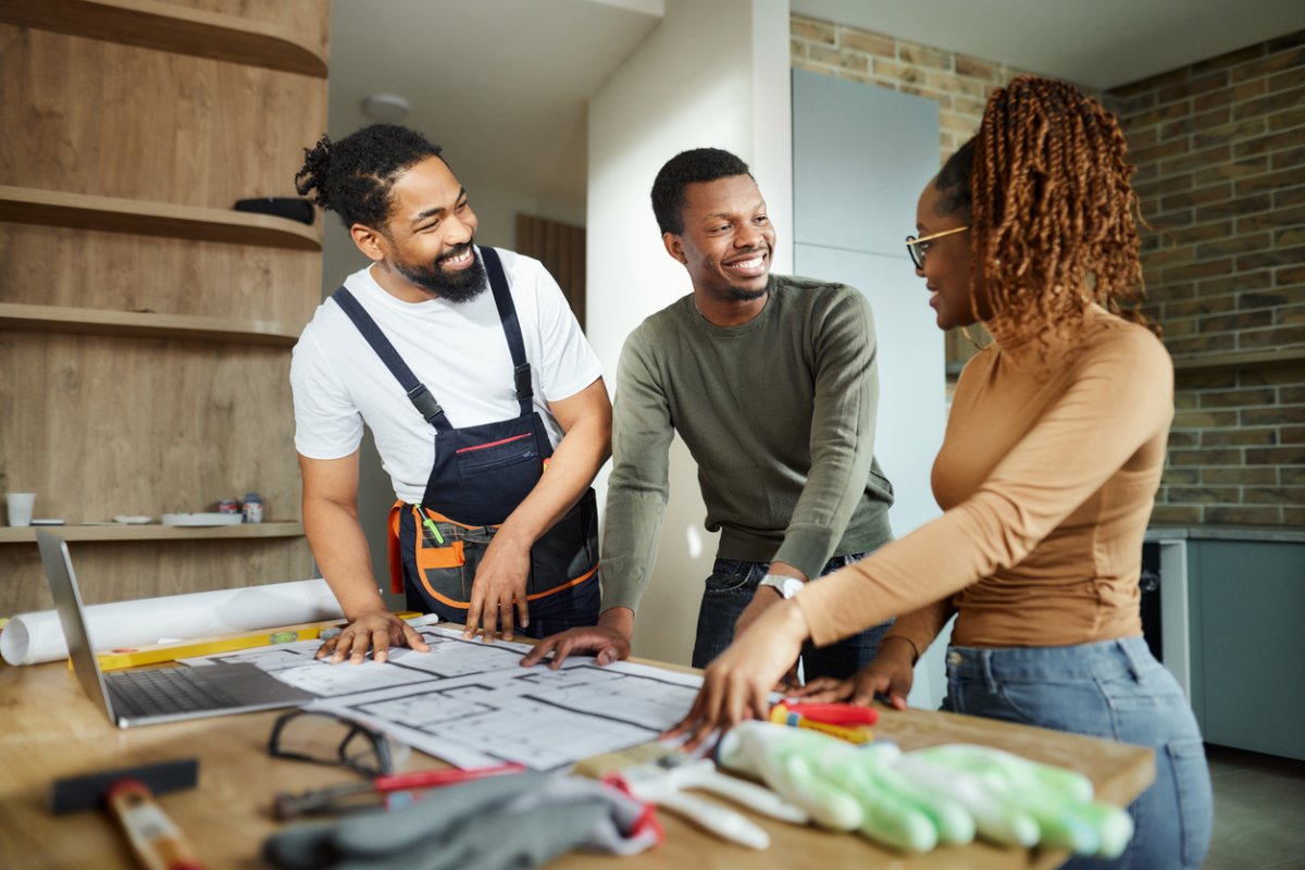 Happy African American couple talking while analyzing blueprints with manual worker in the apartment.