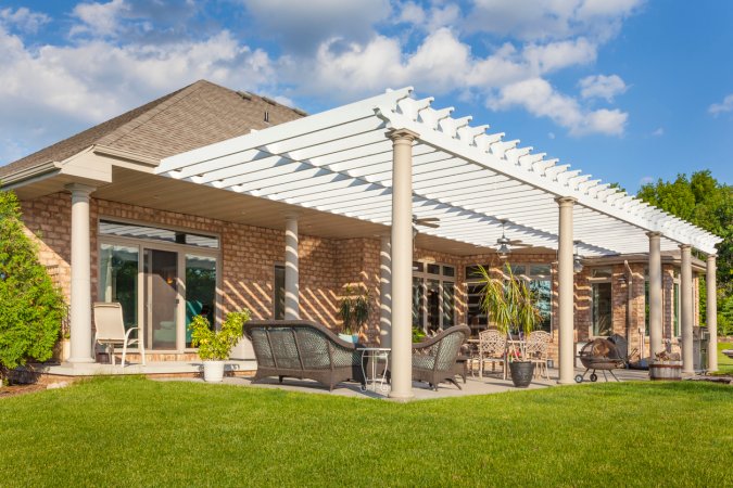 Tan brick ranch style home with white pergola structure in back yard with patio furniture.