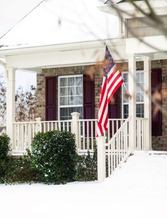 A front porch covered in snow with an American flag hanging out front.