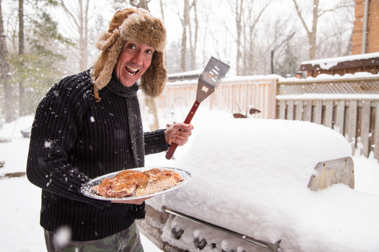 a man approaches his grill with a platter of chicken. He is excited about the prospect of BBQ in spite of the big pile of frozen snow