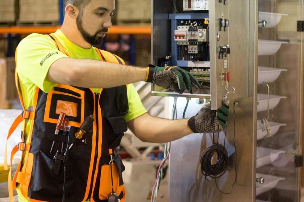 A person wearing a black and orange safety tool vest and gloves while working on an electrical box.