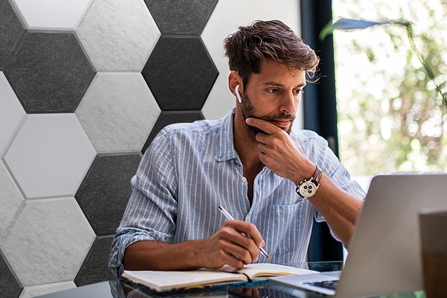 A person working at a computer at their desk with ear buds in and a wall of black and white hexagon soundproofing panels on the wall in the background.