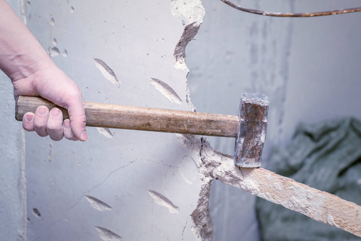 Person holding sledgehammer to smashed wall.