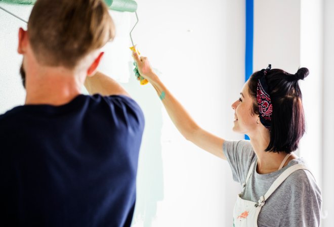 Young couple paint home interior walls with roller brush while smiling.