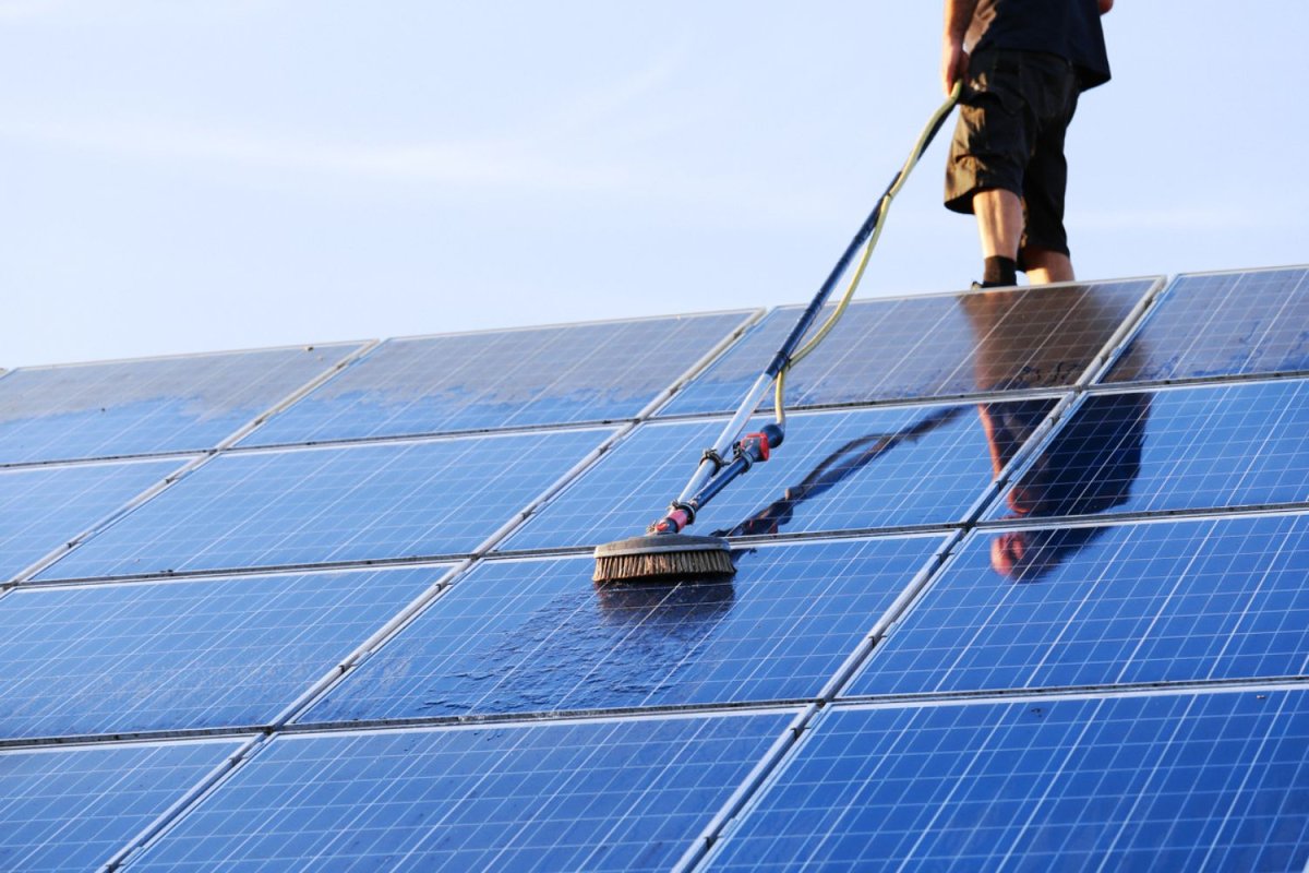 A worker with their bottom half visible cleans a solar panel system on a roof using a brush and a hose.