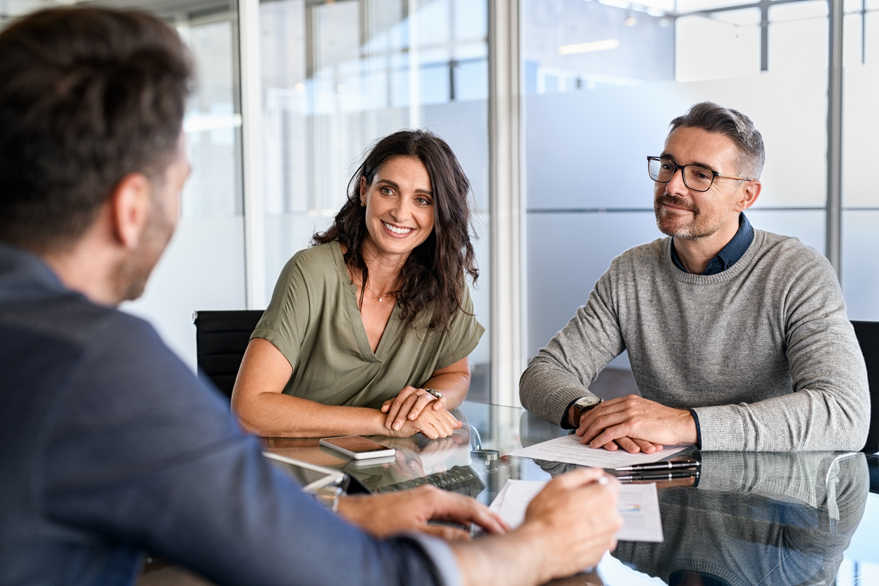 A man and a woman talk to another man sitting across from them at table in an office setting. 