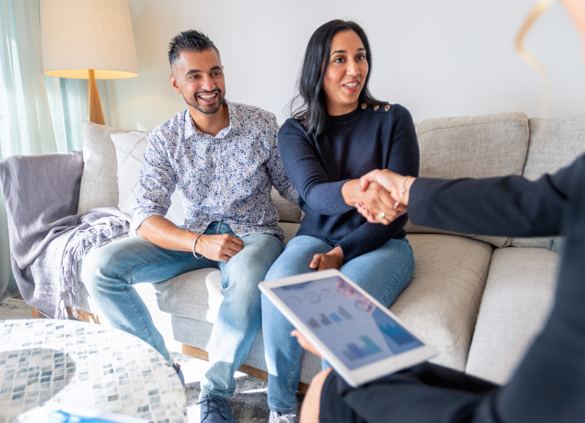 A man and a woman sit on a couch and the woman shakes the hand of someone not in the frame. 