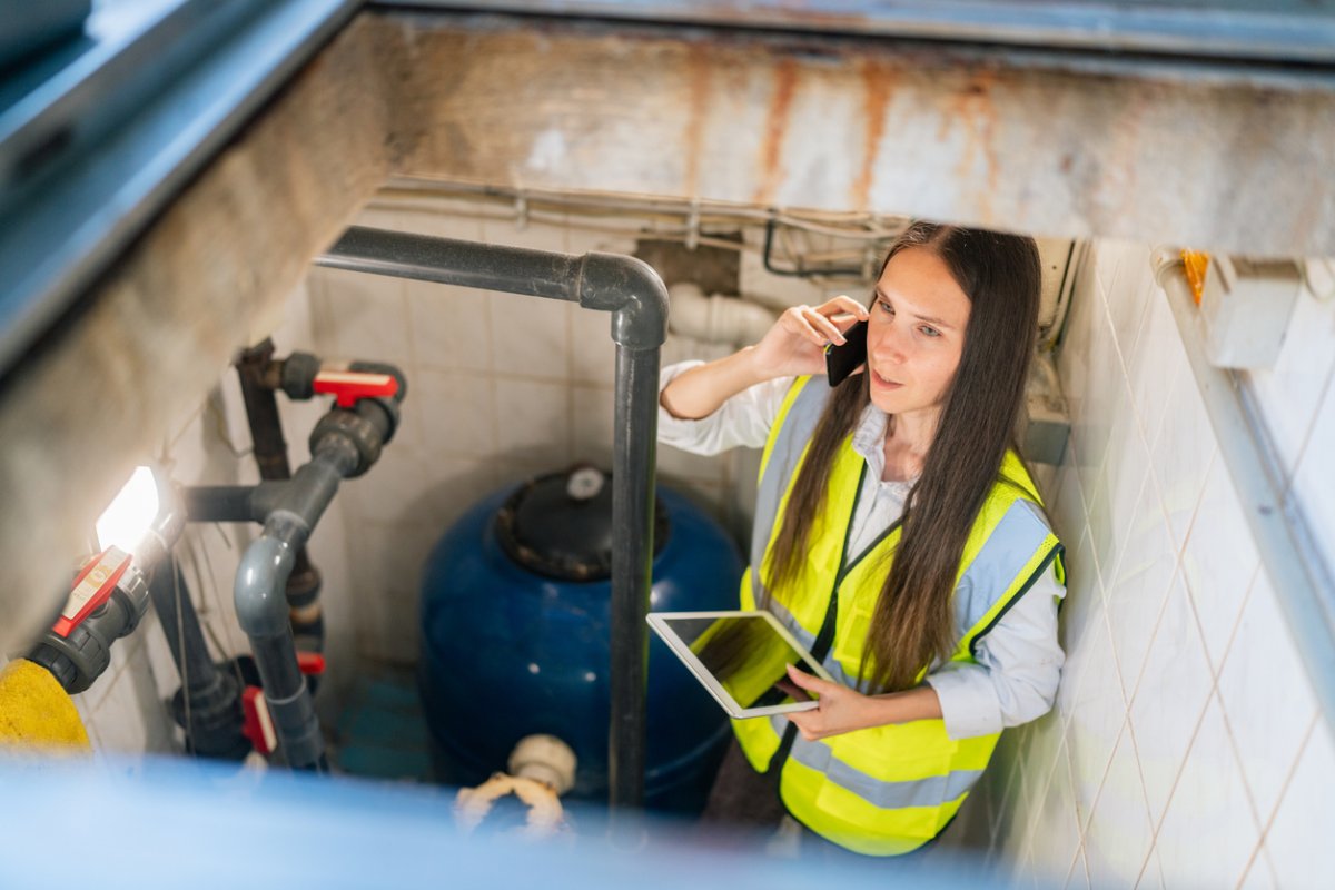 A worker in a yellow vest places a phone call while inspecting pipes.