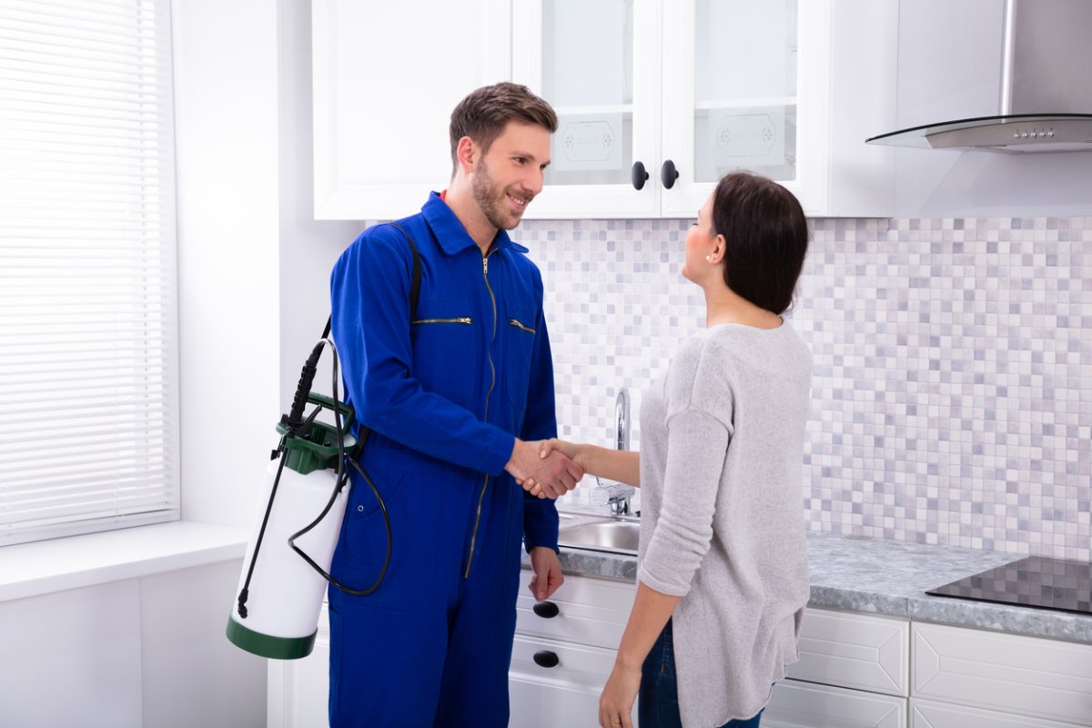 A man in a blue work suit carrying a pest control tool shakes the hand of a woman.