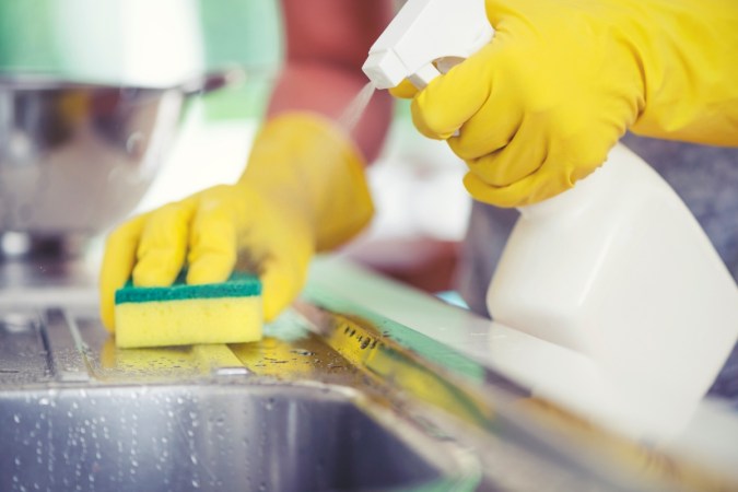 Person cleaning kitchen sink with sponge and spray bottle.