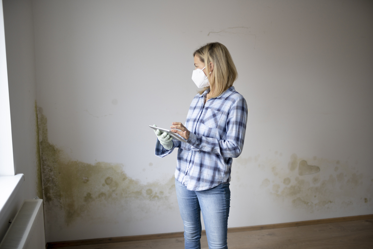 A-person-stands-in-front-of-a-wall-with-mold-along-the-base-while-wearing-a-mask.