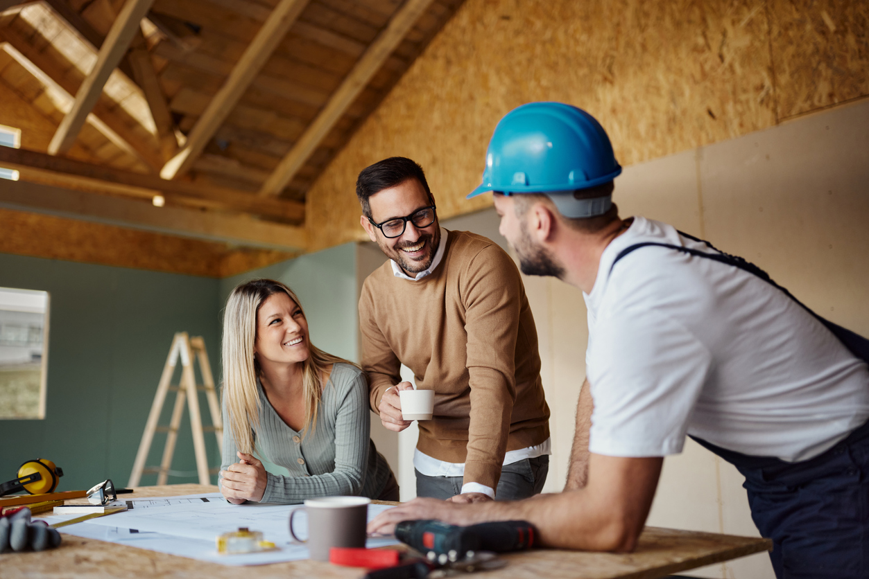 A younger couple speak with construction worker wearing a hardhat in a construction area over plans.