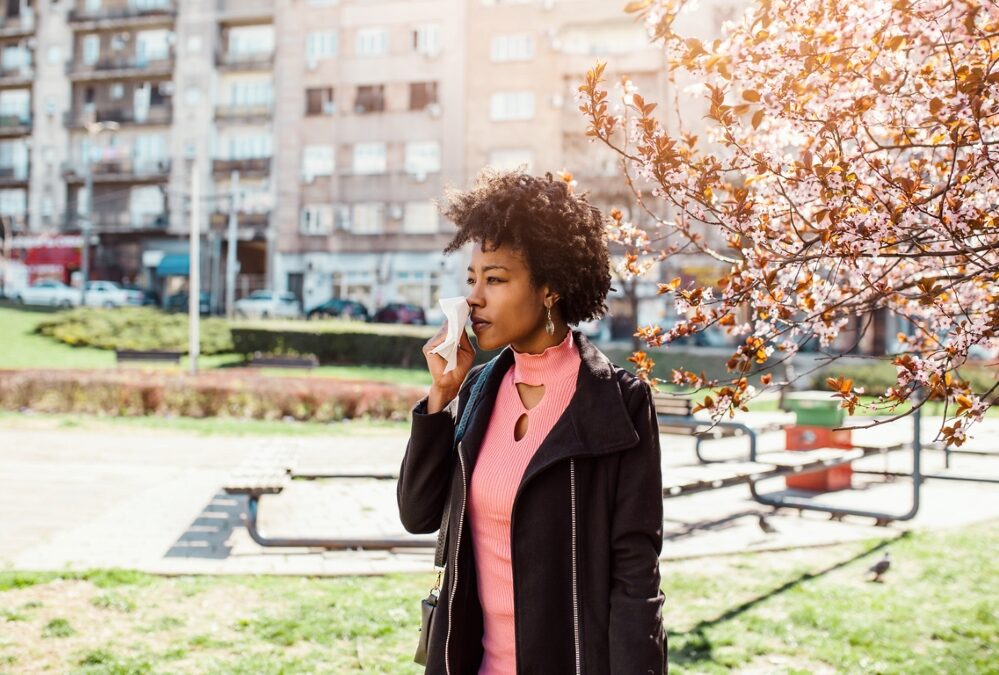 Young woman holding tissue to nose near blooming tree.