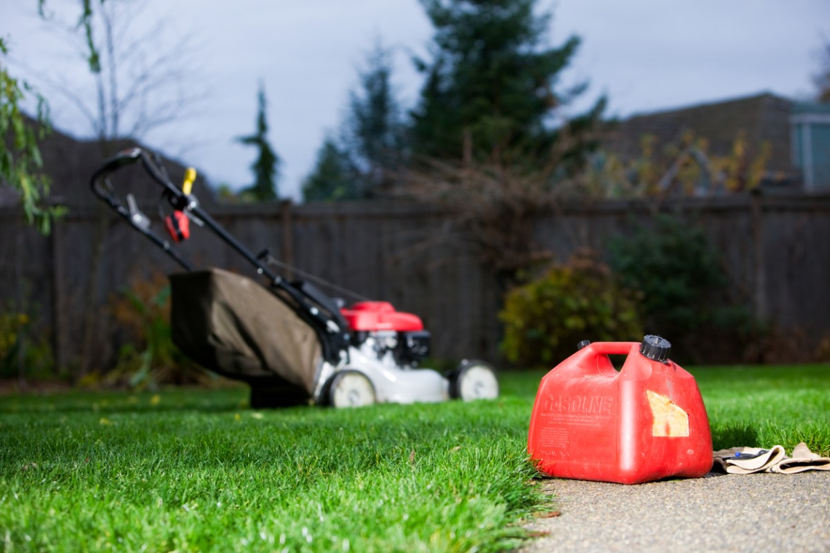 Gasoline Can With Mower In Background. Plastic red gasoline can and leather gloves laying on an aggregate concrete patio. The lawn mower is out of focus in the background. There is a fence in the background. There is also ear protection on the handle of the lawn mower.