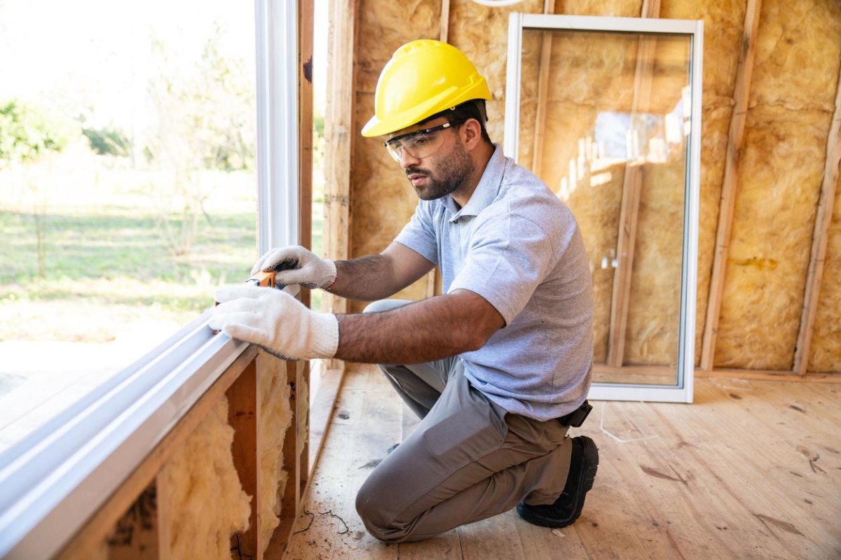 A worker in a yellow hard hat installs a window in a home under construction.