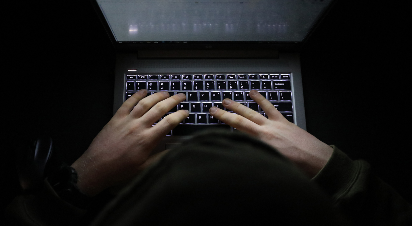 A shrouded person typing on a backlit laptop keyboard in darkness.