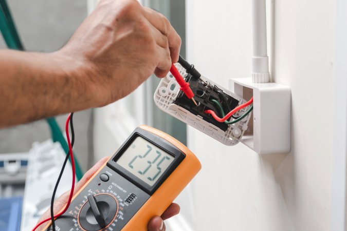 A person uses a voltage meter reading "235" on the back of an unscrewed, but connected, wall outlet.