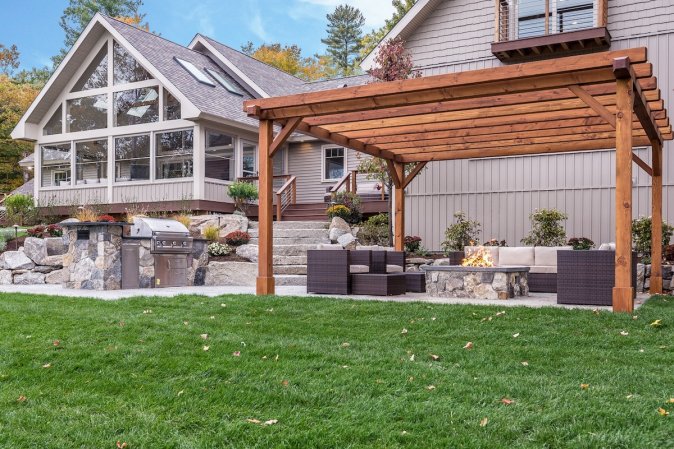 The thoroughly hardscaped backyard of a home, complete with a furnished patio covered by a pergola, a built-in outdoor kitchen, and a hot tub.