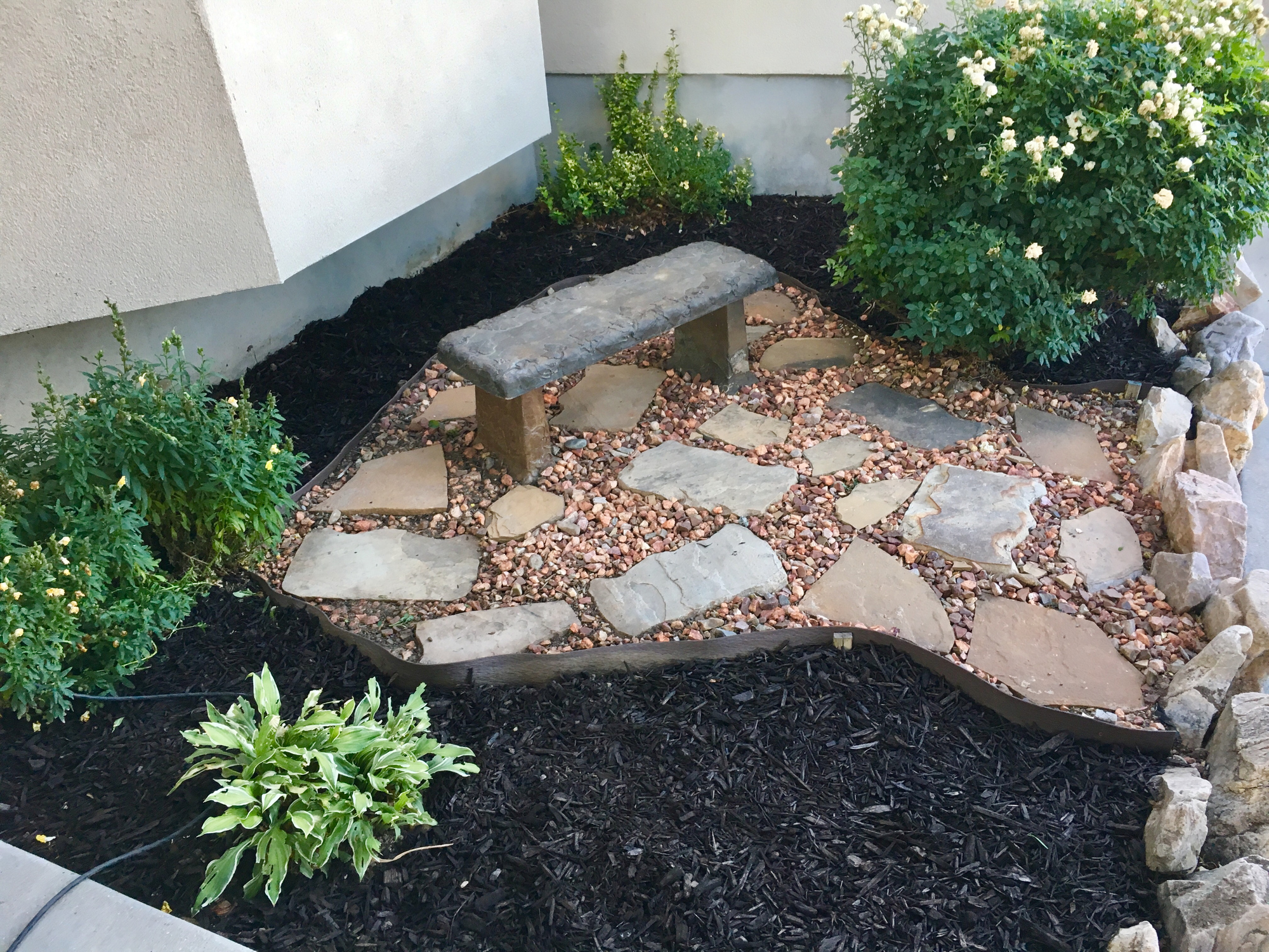 A garden featuring a stone bench, stones, mulch, and flower bushes.