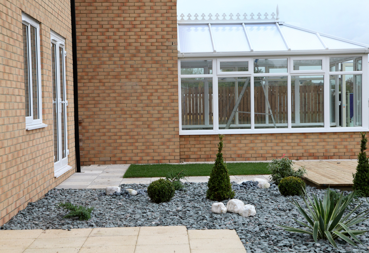 A brick home back yard with a flat stone patio, slate chip rock garden, and ornamental plants and grass features.