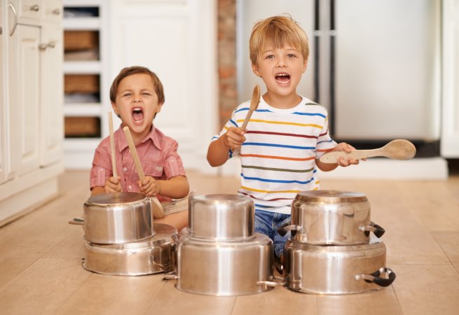 Two young boys sitting on the floor, using kitchen spoons to bang on pots and pans.