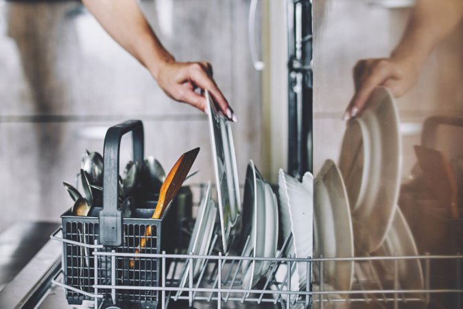 Woman puts a dirty plate in the dishwasher that's loaded with other dirty silverware and dishes.